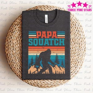 Può includere: Una maglietta grigio scuro con una grafica retrò di un Bigfoot che cammina in un bosco con il testo "PAPA SQUATCH" in rosso, blu e bianco.