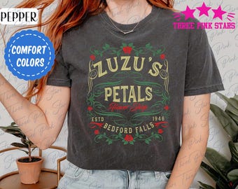 May include: Dark gray t-shirt featuring the text "Zuzu's Petals Flower Shop Bedford Falls 1946" in a vintage design with red roses and green vines. The shirt also has the words "Comfort Colors" in a blue circle.