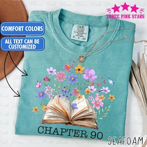 May include: Seafoam green t-shirt with a floral design featuring an open book and colorful flowers. The shirt has the text "CHAPTER 90" and "SEAFOAM" printed on it. The shirt also has the text "COMFORT COLORS" and "ALL TEXT CAN BE CUSTOMIZED" printed on it.