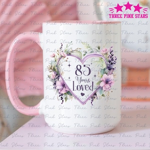 Könnte beinhalten: Eine weiße Keramiktasse mit einem rosa Henkel. Die Tasse zeigt ein violettes Herz mit dem Text "85 Years Loved" umgeben von einem Blumenkranz.