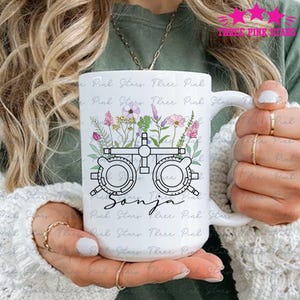 Peut inclure: Une tasse en céramique blanche avec une illustration en noir et blanc d'un tableau d'acuité visuelle avec des fleurs autour. Le nom "Sonja" est écrit sous le tableau d'acuité visuelle.