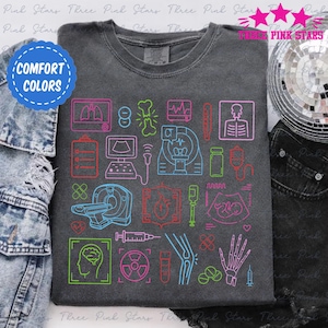 Könnte beinhalten: Ein dunkelgraues T-Shirt mit einem bunten Design aus medizinischen Geräten und Symbolen. Das Design beinhaltet ein Stethoskop, ein Herz, ein Gehirn, einen Knochen, eine Spritze, eine Pille, ein Thermometer, einen CT-Scanner, einen Ultraschall und ein Röntgenbild. Das Design ist in Neonfarben umrandet.