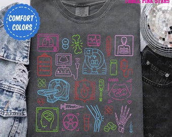 Puede incluir: Una camiseta gris oscuro con un diseño colorido de equipos y símbolos médicos. El diseño incluye un estetoscopio, un corazón, un cerebro, un hueso, una jeringa, una píldora, un termómetro, una tomografía computarizada, una ecografía y una radiografía. El diseño está delineado en colores neón.