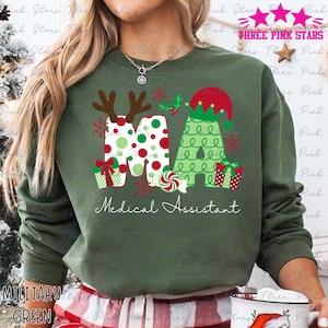 Op de afbeelding: Militairgroen sweatshirt met een kerstthema. Het ontwerp toont de letters "MA" versierd met rendiergeweien, een elfenmuts en vakantie-elementen. De woorden "Medical Assistant" staan eronder gedrukt.