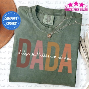 Könnte beinhalten: Ein grünes T-Shirt mit dem Wort "DADA" in braunen, rosa und orangen Buchstaben. Die Namen "Lily", "Kellie" und "Liam" sind in einer kleineren Schrift unter dem Wort "DADA" mit Herzen zwischen den Namen geschrieben.