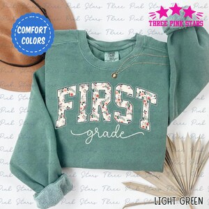 May include: Light green sweatshirt with the words "FIRST grade" in a floral design. The sweatshirt has a crew neck and long sleeves. The words "COMFORT COLORS" and "THREE PINK STARS" are also visible.