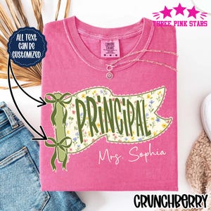 May include: A pink short-sleeved t-shirt featuring a graphic of a flag on a pole with two green bows. The flag has a floral pattern and the text "PRINCIPAL" in green, with "Mrs. Sophia" written below. A dark blue circle states "ALL TEXT CAN BE CUSTOMIZED."