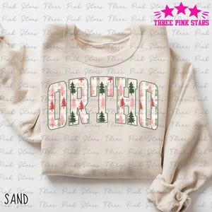 May include: A sand-coloured sweatshirt featuring the word "ORTHO" in large, block letters. The letters are filled with a plaid pattern and Christmas tree graphics in green, pink, and white. The text "THREE PINK STARS" is at the top.