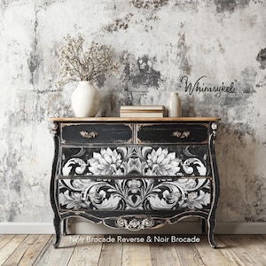 May include: A black wooden chest of drawers featuring a distressed finish and white floral brocade patterns on the lower drawers. The piece has two top drawers with ornate metal pulls, curved legs, and a natural wood top surface with a white vase.