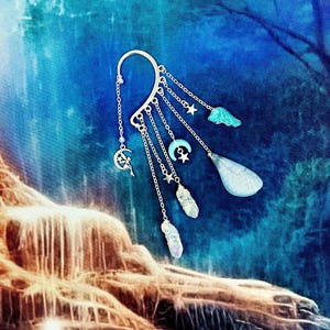 May include: Silver ear cuff with dangling charms, including stars, moons, crystals, and a blue butterfly. The cuff is set against a blue and brown background.