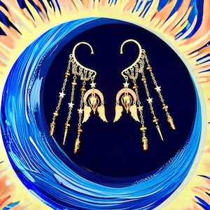 May include: A pair of gold ear cuffs with dangling charms. The charms include stars, beads, and small wings. The cuffs are displayed against a blue and yellow background.