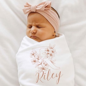 May include: A newborn baby wrapped in a white swaddle blanket, with a matching pink bow headband. The blanket features a floral design with the letter "R" and the name "Riley" in a delicate, light brown color.