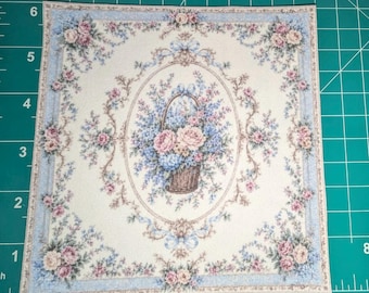 May include: A square fabric patch featuring a central oval frame containing a basket of pink and blue flowers. The border is decorated with a light blue and cream floral pattern. The patch is placed on a green cutting mat with inch markings.