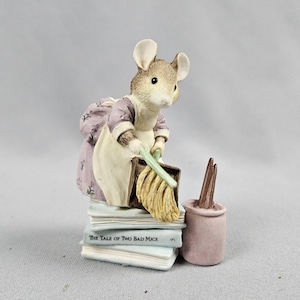 Op de afbeelding: Een beeldje van een muis in een lavendelkleurige jurk met een wit schort, die een gele dweil en een stoffer en blik vasthoudt. De muis staat op een stapel boeken, waarvan er één "The Tale of Two Bad Mice" heet, naast een kleine roze emmer.