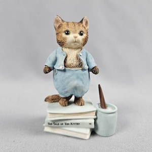 Könnte beinhalten: Eine braune getigerte Katzenfigur in einer hellblauen Jacke und passender Hose, die auf einem Stapel weißer Bücher steht. Das oberste Buch ist deutlich mit dem Text The Tale of Tom Kitten beschriftet. Ein kleiner hellblauer Topf mit einem braunen Werkzeug steht daneben.