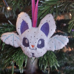 Puede incluir: Adorno navide&ntilde;o de murci&eacute;lago blanco de peluche con orejas y ojos morados, y detalles de alas azul claro. El murci&eacute;lago tiene peque&ntilde;os adornos plateados y una cinta rosa para colgar.