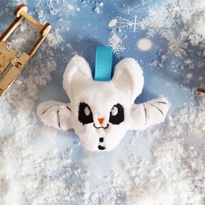 Puede incluir: Adorno de murci&eacute;lago de peluche blanco con ojos negros, una peque&ntilde;a nariz naranja y una boca de bot&oacute;n negra. El murci&eacute;lago tiene alas extendidas y una cinta azul claro para colgar. Fondo con copos de nieve.