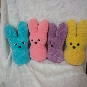 May include: Four plush bunny toys in a row, each with a different color: blue, pink, purple, and yellow. Each bunny has black button eyes and a small, black stitched mouth. The bunnies are sitting on a white, textured surface.