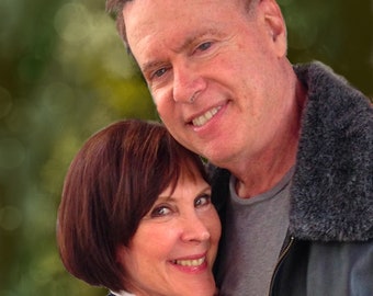 May include: A couple smiling at the camera. The man is wearing a brown leather jacket with a fur collar and a grey t-shirt. The woman is wearing a blue sweater and has her hand on the man's shoulder.