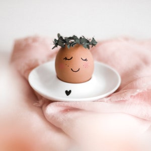 Könnte beinhalten: Ein braunes Ei mit einem gemalten Smiley-Gesicht und geschlossenen Augen sitzt in einem weißen Eierbecher mit einem Herz-Design. Das Ei ist mit einer grünen Blattkrone verziert.