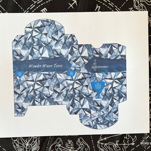May include: A printable template for a box to hold a deck of tarot cards. The box is designed with a blue and white geometric pattern and the text "Wonder Water Tarot" and "Word Witchery".