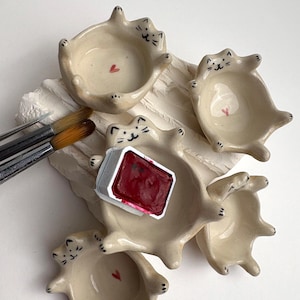 May include: A selection of handmade ceramic cat-shaped paint palettes. Each cream-coloured dish has a painted cat face and a small red heart. A watercolour paint block and paint brushes are also present.
