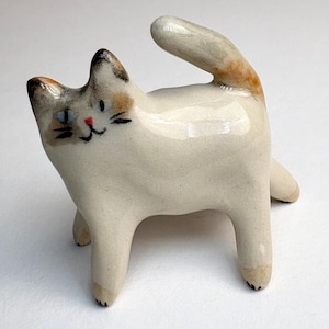 Könnte beinhalten: Keramik-Katzenfigur in spielerischer Pose. Die Katze ist überwiegend weiß mit schwarzen, braunen und orangefarbenen Markierungen an Kopf und Schwanz. Sie hat blaue Augen und eine rote Nase. Die Figur steht auf vier Beinen, der Schwanz ist nach oben gebogen.