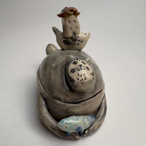 May include: A whimsical ceramic sculpture featuring a gray cat embracing a smaller blue and white fish. A bird-like figure with a mushroom hat sits atop the cat. The sculpture has a matte finish and is set against a white background.