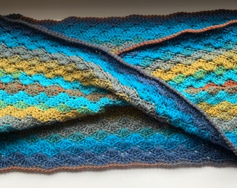 May include: A crocheted blanket in shades of blue, green and brown. The blanket is folded in half and features a textured pattern.