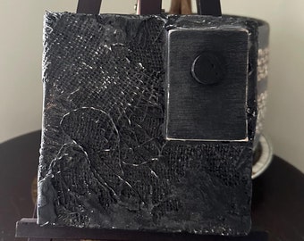 May include: A small black and white textured canvas painting on a wooden easel. The painting has a black square in the upper right corner.