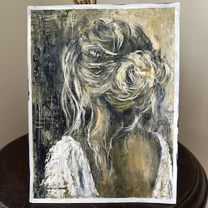 May include: An abstract painting of a person's back and head, with blonde hair styled in a bun. The artwork uses a palette of black, white, and gold, with visible brushstrokes. The painting is framed with a white border.