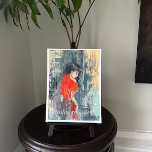 May include: A painting of a woman in a red dress, with a textured background of blue, gray, and white. The painting is on a small easel.