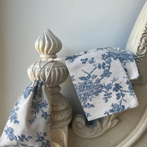 May include: Two small, blue and white floral patterned fabric drawstring bags. The bags are hanging from a white, ornate, wooden post.