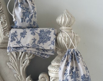 May include: Three small fabric drawstring bags with a blue and white floral pattern. The bags are sitting on a white ornate wooden surface.