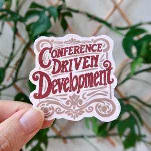 Puede incluir: Una pegatina blanca con un diseño marrón y rojo. La pegatina dice "Conference Driven Development" en una fuente decorativa.