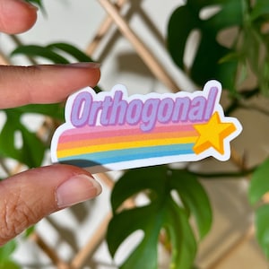 May include: A sticker with the word "Orthogonal" in a purple font, set against a rainbow-colored background with a yellow star. The sticker is held between fingers, with green foliage in the background.