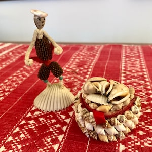 May include: A whimsical figurine made of seashells and twigs, dressed in a red and brown outfit, stands on a seashell base. A small, round basket filled with seashells sits on a red and white patterned surface.