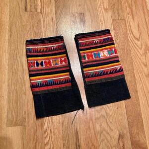 May include: Two black fabric cuffs with colorful embroidered patterns. The cuffs feature horizontal bands of red, yellow, blue, and white, with geometric designs in the center. The cuffs are displayed on a wooden surface.