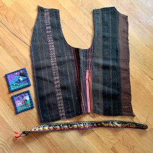 May include: A black and brown striped vest with a red and yellow stripe detail. The vest is made of woven fabric and has a simple design. Two small, colorful, embroidered squares are included, along with a black and gold woven belt.