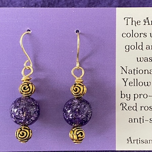 May include: A pair of dangle earrings with gold-colored hooks and decorative elements. Each earring features a gold spiral design above and below a speckled purple bead. The earrings are displayed on a purple card.