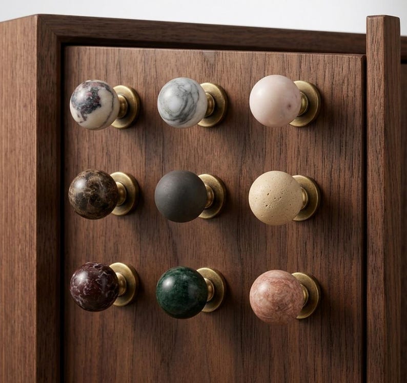 Op de afbeelding: Een donkerhouten kastdeur met negen bolvormige knoppen in een drie-bij-drie raster. Elke knop is gemaakt van een andere steensoort of materiaal, waaronder marmer, graniet en effen kleuren, gemonteerd op ronde messing voetjes.