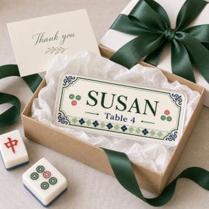 Pode incluir: Um cartão de lugar retangular em acrílico com o nome SUSAN e Table 4 em texto verde escuro. O design inclui um padrão de losangos verde claro, ornamentos de canto azul escuro e pequenos motivos circulares, apresentado numa caixa com fita verde.