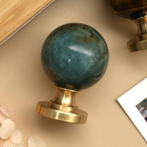 K&ouml;nnte beinhalten: Nahaufnahme von zwei dekorativen Wachssiegelstempeln. Ein Stempel hat eine teal-farbene, kugelf&ouml;rmige Steinspitze mit goldfarbenem Sockel. Der andere Stempel hat eine braune Spitze und einen goldenen Sockel. Die Stempel liegen auf einer beigen Oberfl&auml;che.