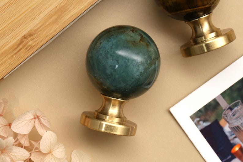 K&ouml;nnte beinhalten: Nahaufnahme von zwei dekorativen Wachssiegelstempeln. Ein Stempel hat eine teal-farbene, kugelf&ouml;rmige Steinspitze mit goldfarbenem Sockel. Der andere Stempel hat eine braune Spitze und einen goldenen Sockel. Die Stempel liegen auf einer beigen Oberfl&auml;che.