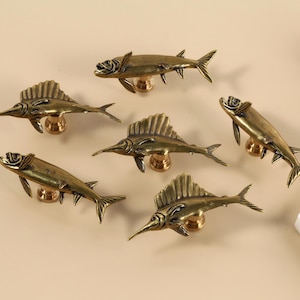 May include: Six bronze-coloured fish-shaped cabinet knobs. The knobs have detailed fish designs with fins and tails, mounted on round brass bases. The knobs are arranged on a neutral background.