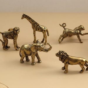 May include: A collection of small, gold-colored animal figurines, including a monkey, giraffe, elephant, cheetah, and lion. The figurines are detailed and appear to be made of metal, with a vintage or antique aesthetic.