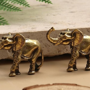 Peut inclure: Deux petites figurines d'éléphants dorés antiques. Les éléphants ont des trompes levées et de grandes oreilles. Ils sont posés sur une surface brun clair, avec un fond en forme de pierre et une fougère en haut à gauche.