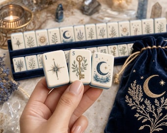 May include: A set of cream-colored tiles featuring botanical and celestial illustrations, including a star, a key with leaves, and a crescent moon. The tiles are displayed in a dark blue velvet rack and a matching drawstring pouch with gold embroidery.
