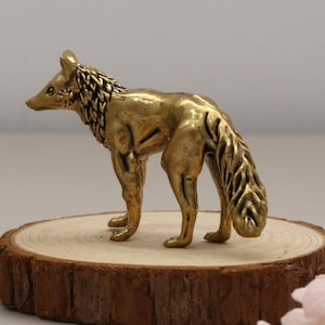May include: A gold-coloured fox figurine stands on a wooden slice. The fox has a detailed mane and tail, with a second fox figurine visible in the background. The figurine is a decorative item, suitable for display.