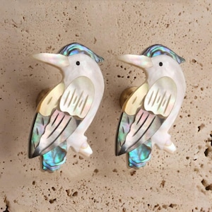 May include: Pair of bird-shaped cabinet knobs. Each knob features a white body with iridescent abalone shell accents on the wings and tail. The knobs have a gold-colored base and a blue iridescent crest.